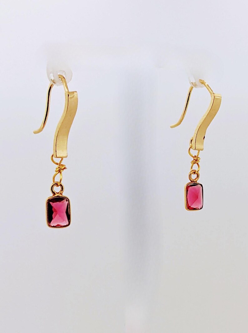 May include: A pair of gold earrings with a dangling pink gemstone. The earrings have a simple, curved design.