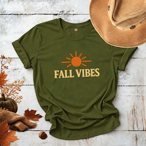 May include: An olive green short-sleeve t-shirt featuring a graphic of an orange sun with rays above the text FALL VIBES in cream-colored letters. The shirt is displayed alongside a tan felt cowboy hat, a small pumpkin, and autumn leaves.