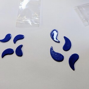 May include: Eight small, glossy, dark blue comma-shaped pieces arranged in two groups of four. Each piece has a smooth, curved surface with a rounded end tapering to a point. The pieces are solid in color and appear to be made of a hard material.