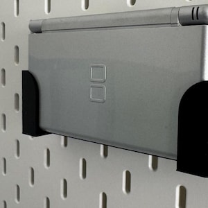 Op de afbeelding: Een zilveren Nintendo DS handheld-spelconsole gemonteerd op een zwarte pegboard-wandbevestiging.