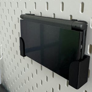 Può includere: Una console per videogiochi portatile Nintendo 3DS nera è montata su una parete di bacheca bianca con un supporto in plastica nera.