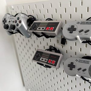 Puede incluir: Cuatro mandos de videojuegos se muestran en una pared blanca de tablero de clavijas. Los mandos son un mando Sony PlayStation gris, un mando Super Nintendo Entertainment System gris, un mando Nintendo Entertainment System gris y un mando Super Nintendo Entertainment System gris. Los mandos están todos cableados y se han montado en la pared con ganchos de plástico blanco.