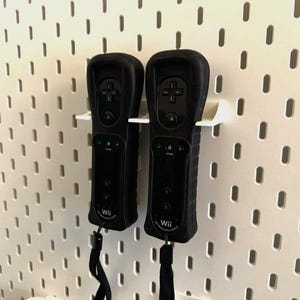 Puede incluir: Dos mandos Wii negros con fundas protectoras montados en una pared perforada blanca. Los mandos tienen botones, un panel direccional y correas para la muñeca. La pared perforada tiene agujeros espaciados.