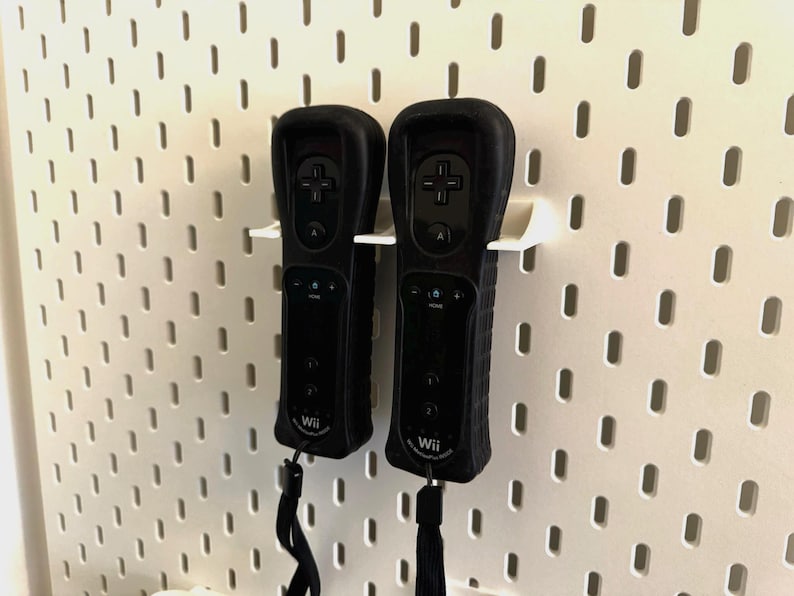 May include: Two black Wii remotes with protective covers are mounted on a white pegboard. The remotes have buttons, a directional pad, and wrist straps. The pegboard has evenly spaced holes.