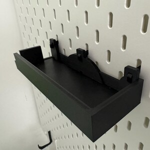 May include: Black shelf with a black metal bracket mounted on a white pegboard wall.