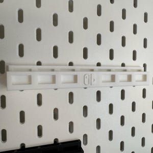K&ouml;nnte beinhalten: Ein wei&szlig;er Kunststoff-Regalaufsatz f&uuml;r eine Lochwand, zur Aufnahme einer Nintendo Switch. Das Regal hat eine rechteckige Form mit einer Reihe quadratischer &Ouml;ffnungen und dem Nintendo Switch-Logo.