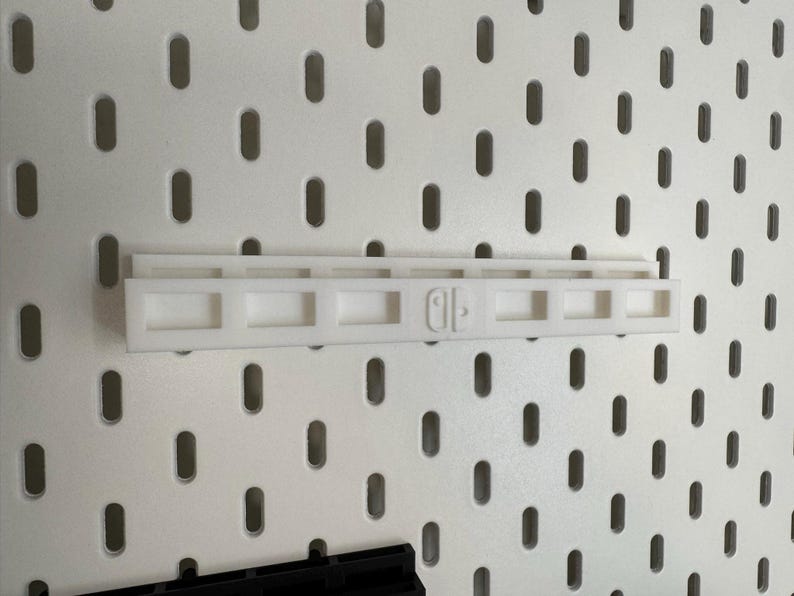 K&ouml;nnte beinhalten: Ein wei&szlig;er Kunststoff-Regalaufsatz f&uuml;r eine Lochwand, zur Aufnahme einer Nintendo Switch. Das Regal hat eine rechteckige Form mit einer Reihe quadratischer &Ouml;ffnungen und dem Nintendo Switch-Logo.