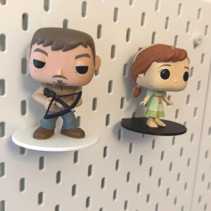Op de afbeelding: Twee pop vinyl figuren op witte pegboard planken. De figuur links is een man met bruin haar en een baard die een kruisboog vasthoudt. De figuur rechts is een vrouw met rood haar die een groene jurk draagt.