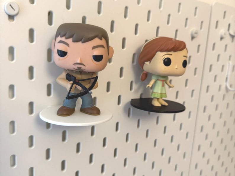 Op de afbeelding: Twee pop vinyl figuren op witte pegboard planken. De figuur links is een man met bruin haar en een baard die een kruisboog vasthoudt. De figuur rechts is een vrouw met rood haar die een groene jurk draagt.