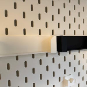 Puede incluir: Dos estantes de almacenamiento montados en la pared. Uno es blanco, el otro negro. Ambos son rectangulares con bordes curvos, dise&ntilde;ados para fijarse a una panel perforado. El panel perforado es blanco con agujeros ovalados espaciados.