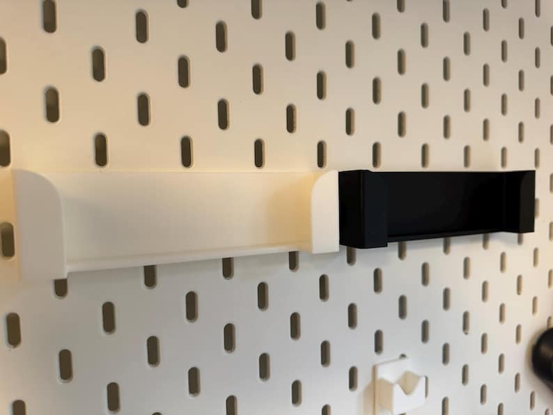 Puede incluir: Dos estantes de almacenamiento montados en la pared. Uno es blanco, el otro negro. Ambos son rectangulares con bordes curvos, dise&ntilde;ados para fijarse a una panel perforado. El panel perforado es blanco con agujeros ovalados espaciados.