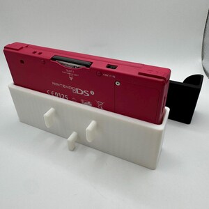 K&ouml;nnte beinhalten: Eine pinkfarbene Nintendo 3DS-Konsole liegt in einem wei&szlig;en 3D-gedruckten Halter mit drei Stiften. Rechts befindet sich ein schwarzer St&auml;nder. Die Konsole ist mit "Nintendo 3DS" und "SLOT 1" beschriftet.