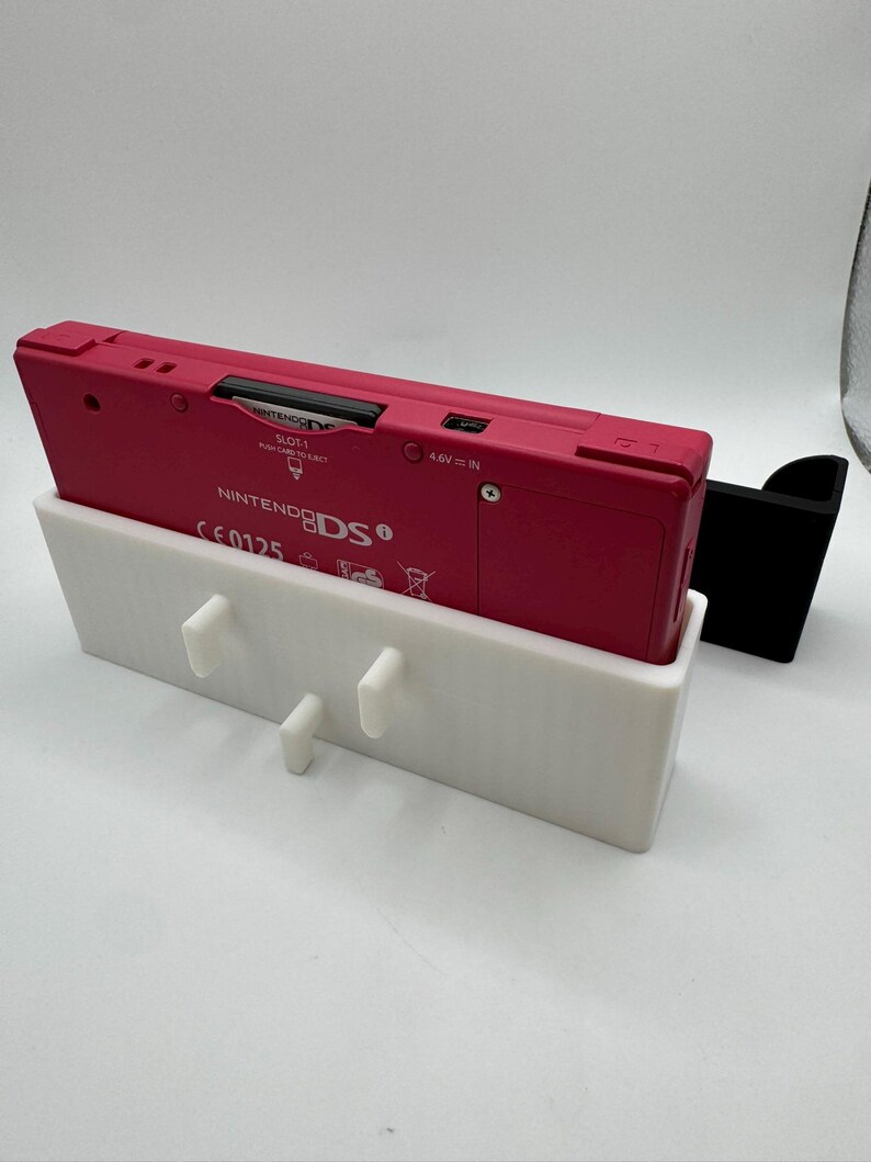 K&ouml;nnte beinhalten: Eine pinkfarbene Nintendo 3DS-Konsole liegt in einem wei&szlig;en 3D-gedruckten Halter mit drei Stiften. Rechts befindet sich ein schwarzer St&auml;nder. Die Konsole ist mit "Nintendo 3DS" und "SLOT 1" beschriftet.