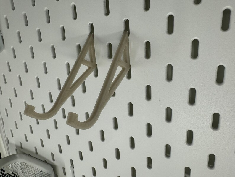 May include: Two beige plastic wall mounted hooks for hanging items on a pegboard.