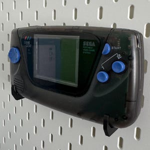 May include: A black Sega Game Gear handheld video game console with blue buttons. The console has a screen displaying a video game. The console is mounted on a white pegboard.