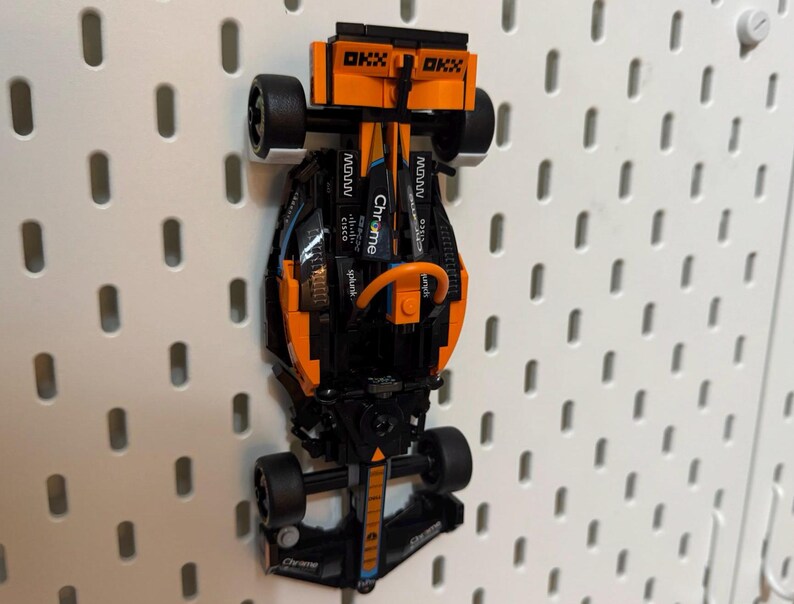 Op de afbeelding: Een zwarte en oranje Lego raceauto met de woorden "Chrome" en "spin" aan de zijkant. De auto hangt aan een prikbord.