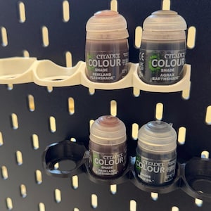 Puede incluir: Un soporte para botes de pintura de pl&aacute;stico blanco y negro con cuatro ranuras para sujetar botes de pintura peque&ntilde;os. El soporte est&aacute; montado en una pared de clavijas negra. Hay cuatro botes de pintura en el soporte. Los botes de pintura est&aacute;n etiquetados como "Citadel Colour" y tienen diferentes tonos de pintura marr&oacute;n y negra.