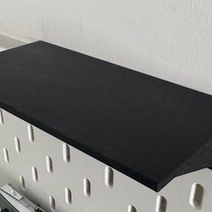 Puede incluir: Una estanter&iacute;a negra de pl&aacute;stico o metal est&aacute; montada en una pared de tablero de clavijas blanco. La estanter&iacute;a est&aacute; ligeramente inclinada hacia abajo y tiene una superficie lisa y plana.