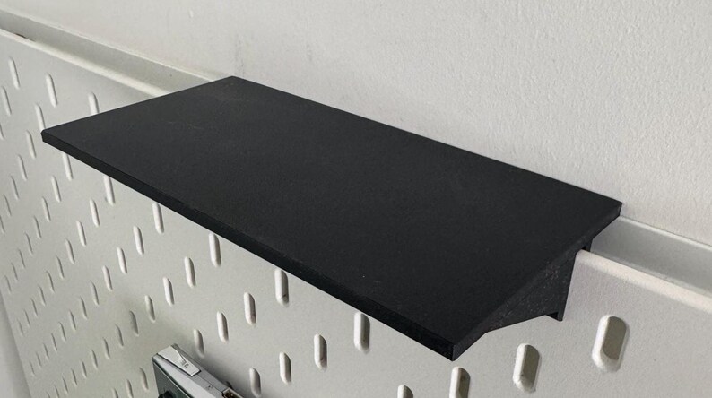 Puede incluir: Una estanter&iacute;a negra de pl&aacute;stico o metal est&aacute; montada en una pared de tablero de clavijas blanco. La estanter&iacute;a est&aacute; ligeramente inclinada hacia abajo y tiene una superficie lisa y plana.