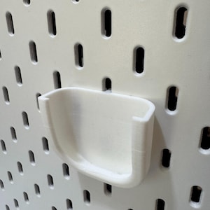 K&ouml;nnte beinhalten: Ein wei&szlig;es, 3D-gedrucktes Lochwandzubeh&ouml;r, ein kleiner, gebogener Halter, ist an einer wei&szlig;en Lochwand befestigt. Der Halter hat ein U-f&ouml;rmiges Design und ist f&uuml;r die L&ouml;cher der Lochwand konzipiert.
