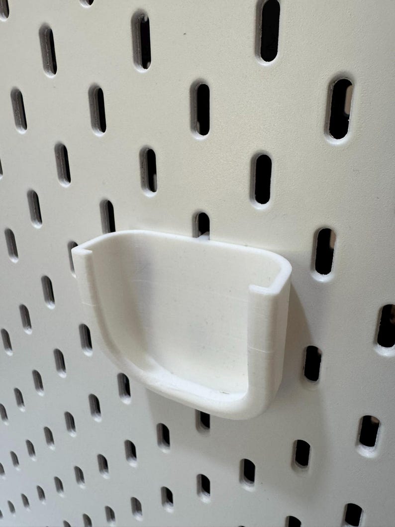 K&ouml;nnte beinhalten: Ein wei&szlig;es, 3D-gedrucktes Lochwandzubeh&ouml;r, ein kleiner, gebogener Halter, ist an einer wei&szlig;en Lochwand befestigt. Der Halter hat ein U-f&ouml;rmiges Design und ist f&uuml;r die L&ouml;cher der Lochwand konzipiert.