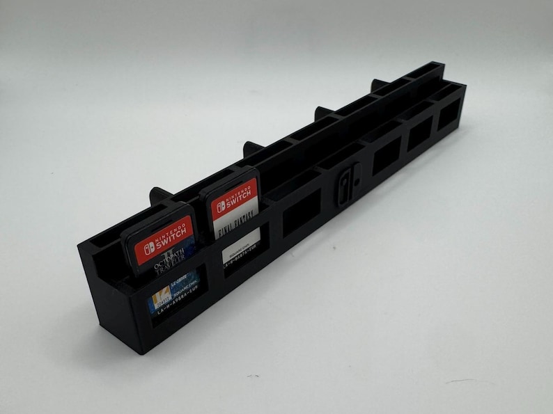 K&ouml;nnte beinhalten: Schwarzer Game-Cartridge-Halter aus Kunststoff f&uuml;r Nintendo Switch. Der Halter hat mehrere Schlitze f&uuml;r Spielkassetten und ein Nintendo Switch-Logo. Mehrere Spielkassetten sind in den Schlitzen sichtbar.