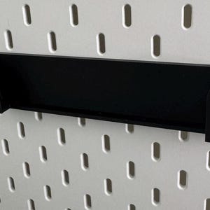 Op de afbeelding: Zwart metalen plank met afgeronde randen, ontworpen om te hangen aan een pegboard muur.
