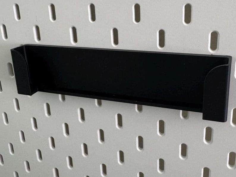 Op de afbeelding: Zwart metalen plank met afgeronde randen, ontworpen om te hangen aan een pegboard muur.