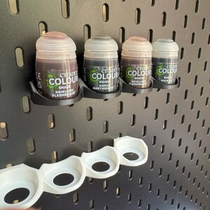 Puede incluir: Cuatro peque&ntilde;os frascos de pintura Citadel Colour, montados en una pared de clavijas negra con un soporte de pl&aacute;stico blanco debajo. Los tonos de pintura son Reikland Fleshshade, Agrax Earthshade, Reikland Fleshshade y Nuln Oil.
