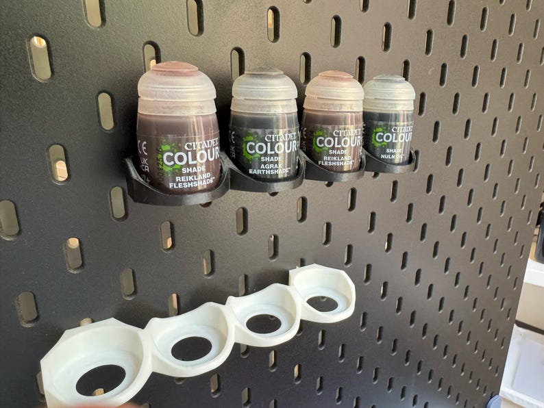 Puede incluir: Cuatro peque&ntilde;os frascos de pintura Citadel Colour, montados en una pared de clavijas negra con un soporte de pl&aacute;stico blanco debajo. Los tonos de pintura son Reikland Fleshshade, Agrax Earthshade, Reikland Fleshshade y Nuln Oil.