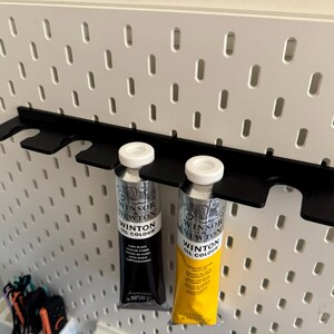 May include: A black metal paint tube holder attached to a white pegboard. Two tubes of Winsor & Newton Winton Oil Colour are in the holder. One tube is black with white text, and the other is yellow with black text.