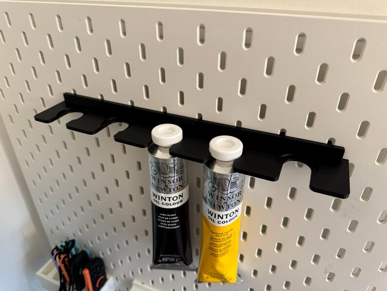 May include: A black metal paint tube holder attached to a white pegboard. Two tubes of Winsor & Newton Winton Oil Colour are in the holder. One tube is black with white text, and the other is yellow with black text.