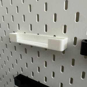 May include: A white, rectangular pegboard shelf is mounted on a white pegboard. The shelf is made of a solid, opaque material. The pegboard has evenly spaced, vertical slots. A black pegboard accessory is visible.