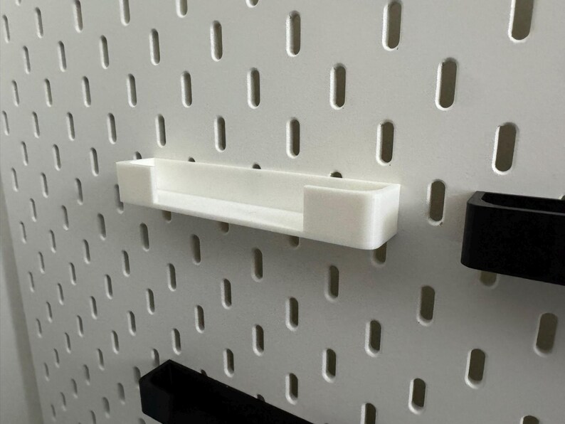 May include: A white, rectangular pegboard shelf is mounted on a white pegboard. The shelf is made of a solid, opaque material. The pegboard has evenly spaced, vertical slots. A black pegboard accessory is visible.