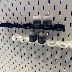 以下が含まれることがあります： 白のペグボードに、黒と白の塗料ポットホルダーが2列並んでいます。ホルダーには、4つの小さな塗料ボトルが入れられており、ボトルには「Citadel Colour」とラベルが貼られ、茶色とグレーの異なる色合いになっています。
