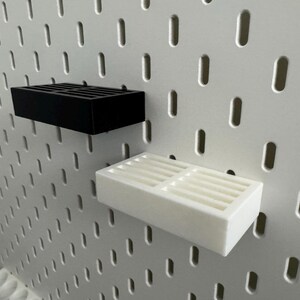 Op de afbeelding: Twee rechthoekige planken, &eacute;&eacute;n zwart en &eacute;&eacute;n wit, zijn gemonteerd op een wit pegboard. Elke plank heeft een roosterachtig ontwerp op het bovenoppervlak. Het pegboard heeft gelijkmatig verdeelde, verticale ovale gaten.