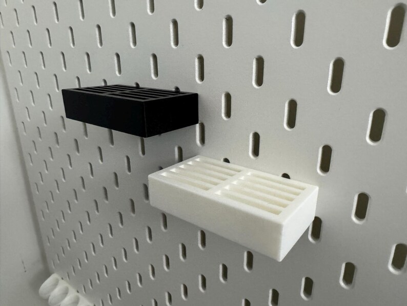 Op de afbeelding: Twee rechthoekige planken, &eacute;&eacute;n zwart en &eacute;&eacute;n wit, zijn gemonteerd op een wit pegboard. Elke plank heeft een roosterachtig ontwerp op het bovenoppervlak. Het pegboard heeft gelijkmatig verdeelde, verticale ovale gaten.