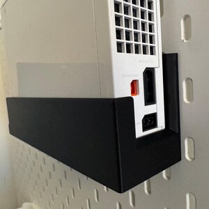 Peut inclure: Une console Nintendo Wii blanche mont&eacute;e sur un support en m&eacute;tal noir. La console est positionn&eacute;e verticalement, montrant les ports et les ouvertures. Le support est fix&eacute; &agrave; un mur perfor&eacute; blanc.