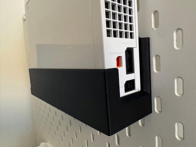 Peut inclure: Une console Nintendo Wii blanche mont&eacute;e sur un support en m&eacute;tal noir. La console est positionn&eacute;e verticalement, montrant les ports et les ouvertures. Le support est fix&eacute; &agrave; un mur perfor&eacute; blanc.