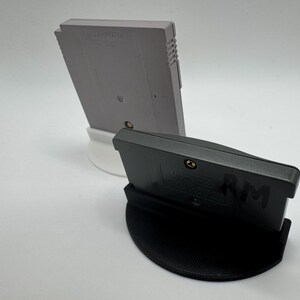 May include: Two video game cartridges, one gray and one black, are displayed on white and black stands. The gray cartridge is upright, while the black one is angled forward. The stands provide a clean, minimalist presentation for the retro gaming items.