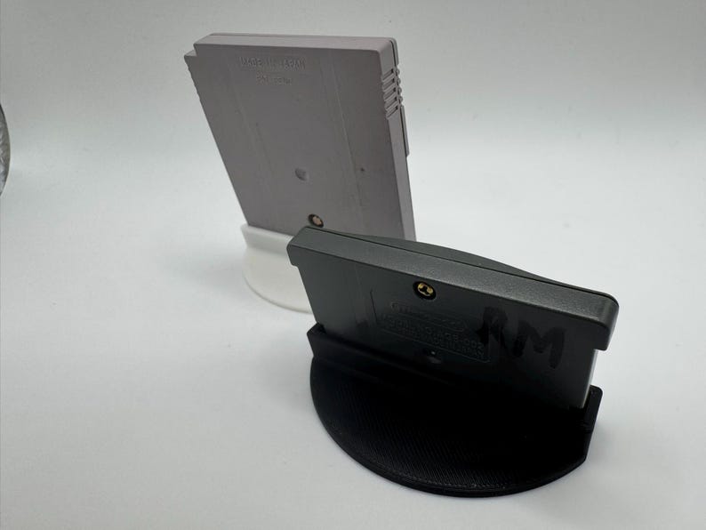 May include: Two video game cartridges, one gray and one black, are displayed on white and black stands. The gray cartridge is upright, while the black one is angled forward. The stands provide a clean, minimalist presentation for the retro gaming items.