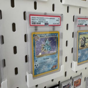 Peut inclure: Une carte Pok&eacute;mon Misty's Seadra class&eacute;e, une carte holographique de pr&eacute;-sortie de 2000. La carte est dans un &eacute;tui de protection transparent et expos&eacute;e sur un panneau perfor&eacute; blanc. D'autres cartes Pok&eacute;mon et cartouches de jeu sont &eacute;galement visibles.