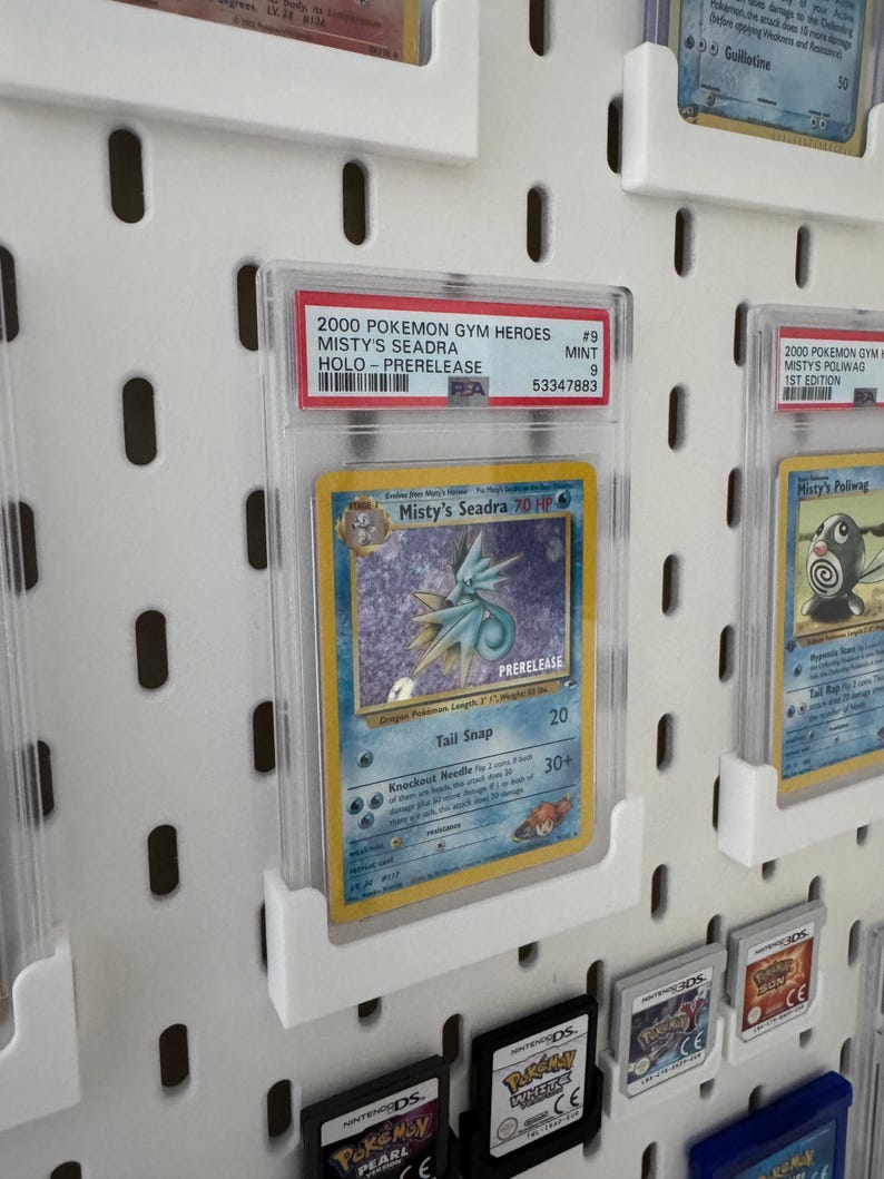 Peut inclure: Une carte Pok&eacute;mon Misty's Seadra class&eacute;e, une carte holographique de pr&eacute;-sortie de 2000. La carte est dans un &eacute;tui de protection transparent et expos&eacute;e sur un panneau perfor&eacute; blanc. D'autres cartes Pok&eacute;mon et cartouches de jeu sont &eacute;galement visibles.