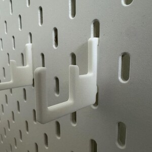 May include: Close-up of a white pegboard with two white hooks inserted. The pegboard has evenly spaced, oblong holes. The hooks are designed to hold items on the pegboard. The hooks are made of a smooth, solid material.