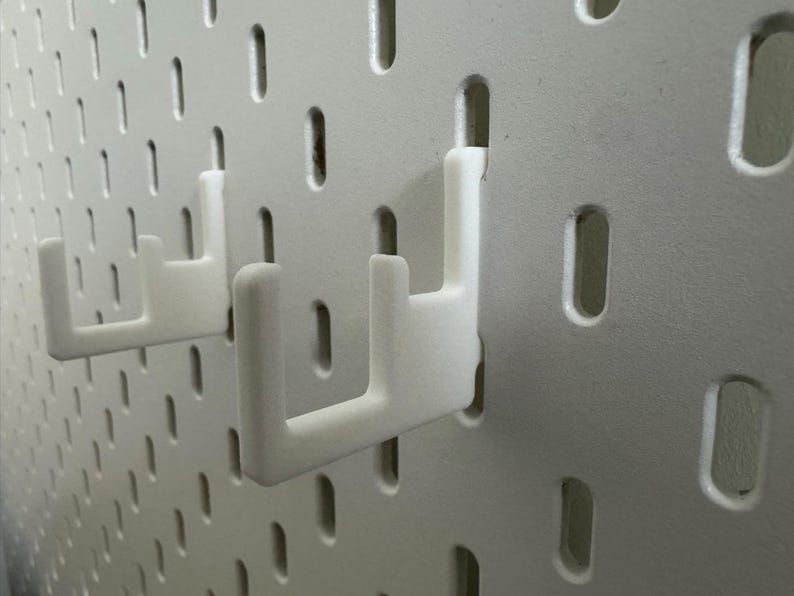 May include: Close-up of a white pegboard with two white hooks inserted. The pegboard has evenly spaced, oblong holes. The hooks are designed to hold items on the pegboard. The hooks are made of a smooth, solid material.