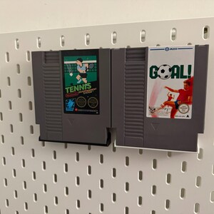 Pode incluir: Dois cartuchos de videogame retr&ocirc; s&atilde;o exibidos. Um &eacute; rotulado "Tennis" com um gr&aacute;fico de jogador, e o outro diz "Goal!" com um jogador de futebol. Ambos s&atilde;o cinza e montados em uma parede perfurada branca.