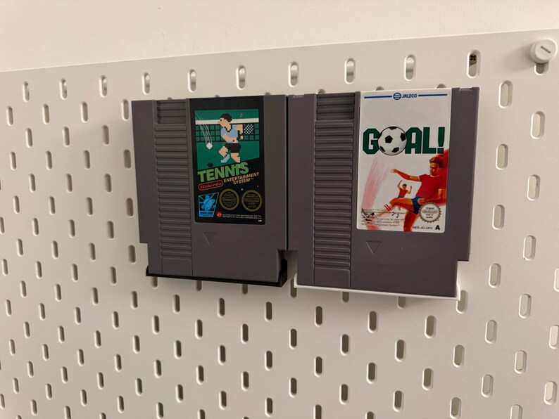 Pode incluir: Dois cartuchos de videogame retr&ocirc; s&atilde;o exibidos. Um &eacute; rotulado "Tennis" com um gr&aacute;fico de jogador, e o outro diz "Goal!" com um jogador de futebol. Ambos s&atilde;o cinza e montados em uma parede perfurada branca.