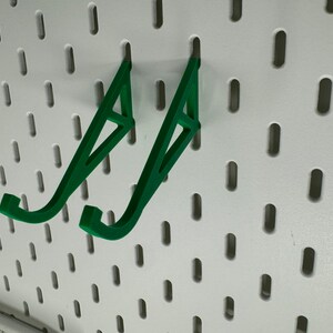 May include: Two green plastic wall mounted hooks for hanging items on a pegboard.