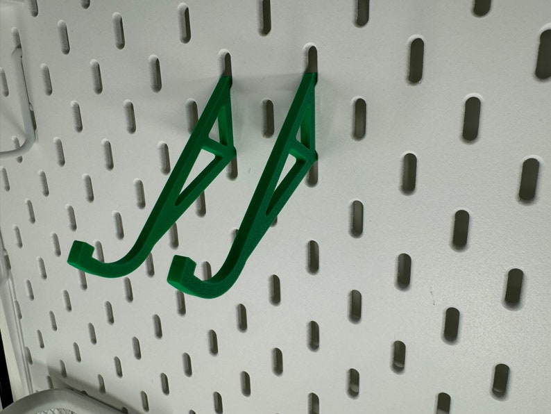 May include: Two green plastic wall mounted hooks for hanging items on a pegboard.
