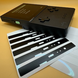 May include: A black handheld gaming console with a green button and a screen displaying the text "Library, Cart, Memories, Tools, Settings". The console is accompanied by a sheet of black and white stickers.